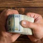 Hands flipping through a stack of one-hundred-dollar bills above a brown wooden table