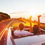 Couple in a convertible driving toward a summer sunset on a paved road