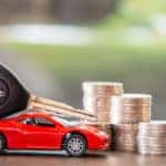 Red toy car with a car key on top sitting next to stacks of change on a brown table