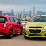 One red and one green 2015 Chevrolet Spark hatchbacks parked in front of water, boats, and tall city buildings