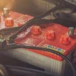 Hands placing a red and white vehicle battery under the open hood of a vehicle