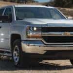 White 2016 Chevrolet Silverado pickup truck parked on flat brown ground in front of a brown fence, a hill, and green trees