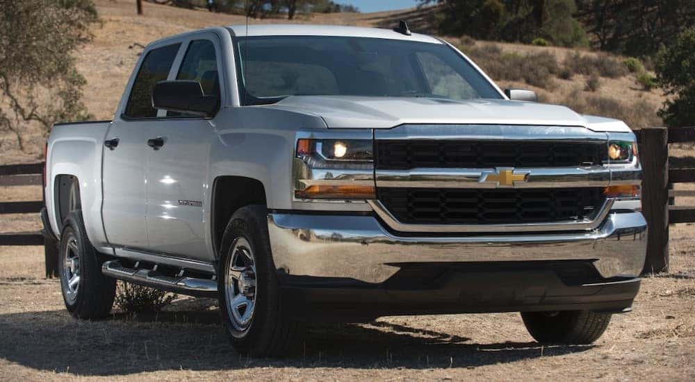 White 2016 Chevrolet Silverado pickup truck parked on flat brown ground in front of a brown fence, a hill, and green trees