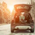 Woman in winter clothing sitting in the open trunk of a vehicle in the middle of a snowy road