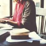 Woman in a pink shirt and grey cardigan sitting at a desk with a laptop and notebooks