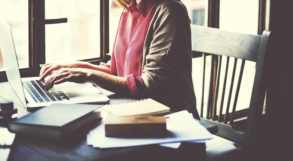 Woman in a pink shirt and grey cardigan sitting at a desk with a laptop and notebooks