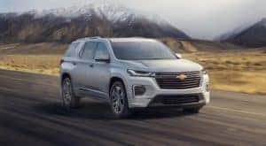A white 2022 Chevy Traverse is driving on a highway away from snow-covered mountains.