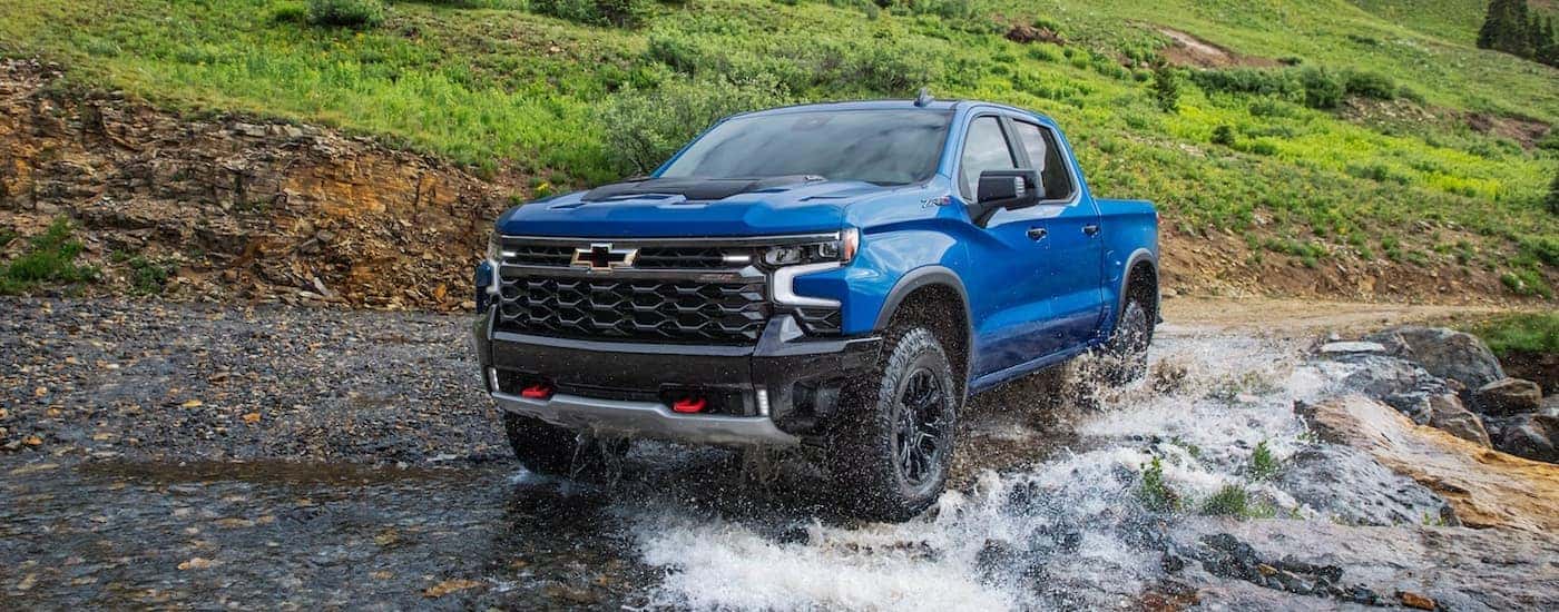 A blue 2022 Chevy Silverado 1500 ZR2 is shown off-roading through a river after leaving an Albany car dealership.