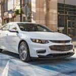A white 2016 Chevy Malibu is driving through a city intersection after leaving a used car dealer in Albany.