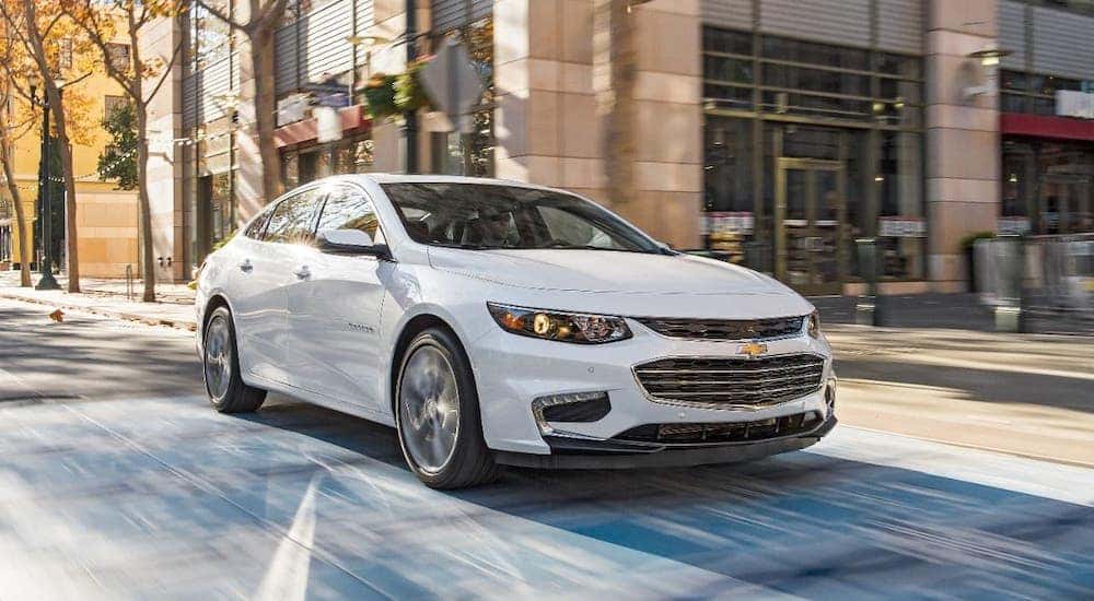 A white 2016 Chevy Malibu is driving through a city intersection after leaving a used car dealer in Albany.