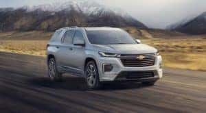 A white 2022 Chevy Traverse High Country is driving in front of snow-covered mountains.