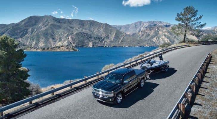 A black 2018 Chevy Silverado 1500 High Country is shown from a high angle towing a boat over a road next to lake.