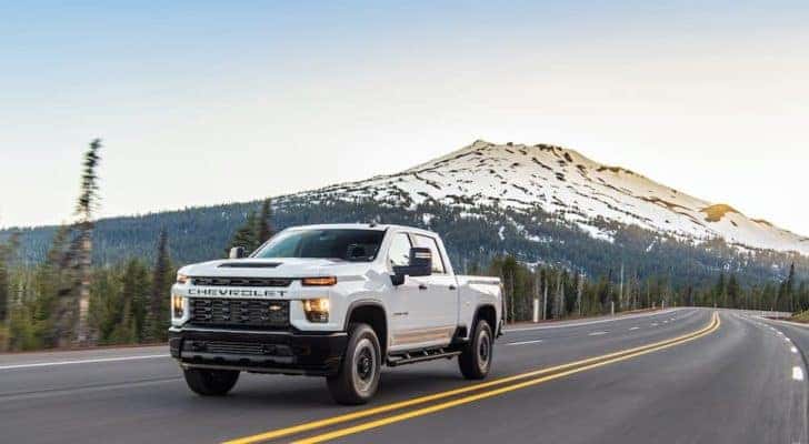 A white 2021 Chevy Silverado 2500 HD is shown driving down the road with mountains in the distance.