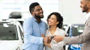 A couple is standing in a Chevy Tahoe dealership as the salesman passes them a set of keys.