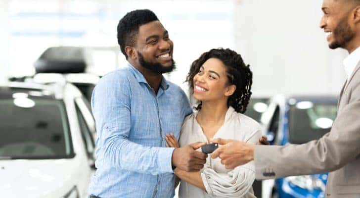 A couple is standing in a Chevy Tahoe dealership as the salesman passes them a set of keys.