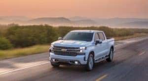 A white 2021 Chevy Silverado High Country is driving down a road against a pastel sky.