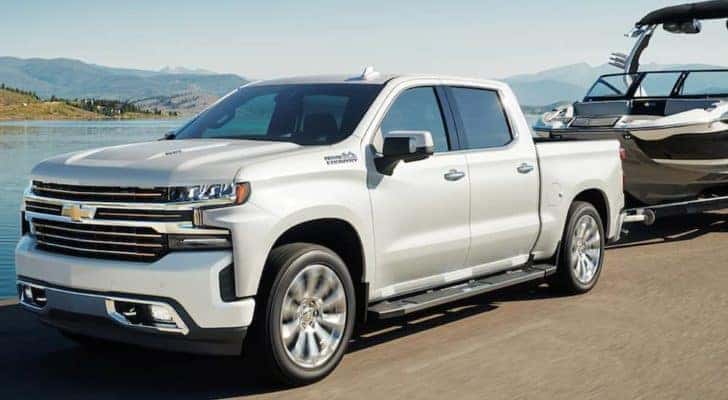 A white 2022 Chevy Silverado 1500 High Country is shown towing a boat past a lake.