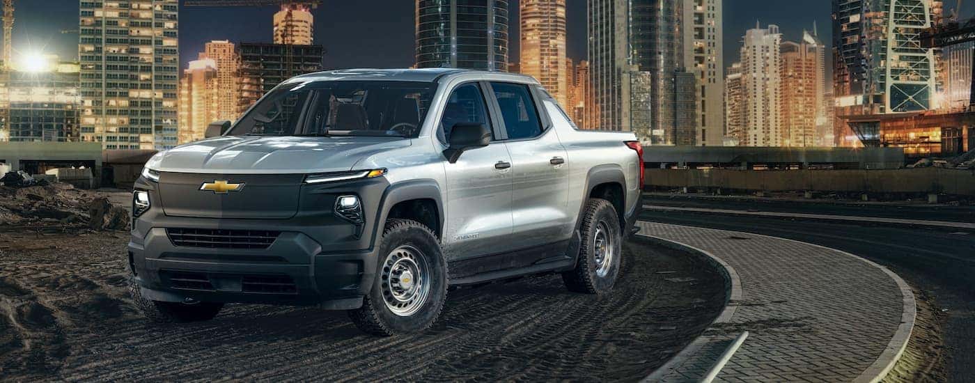 A silver 2024 Chevy Silverado EV is shown parked near a city at night.