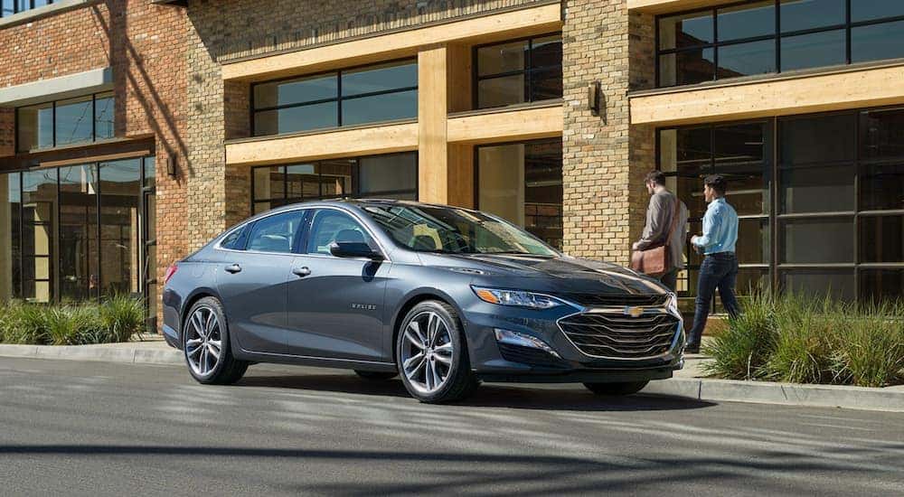 A grey 2020 Chevy Malibu is shown parked outside of a modern office building.