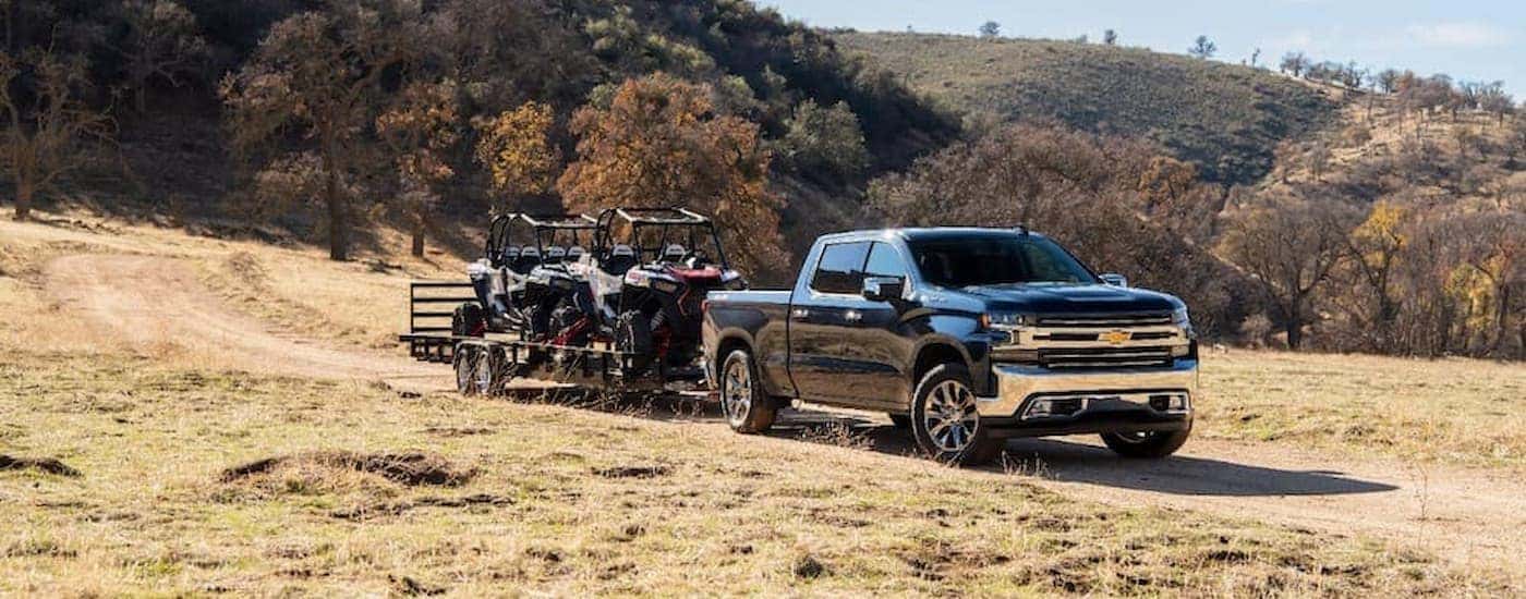 A blue 2020 Chevy Silverado 1500 is shown towing an ATV on a dirt road.