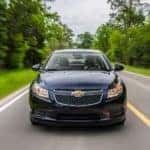 A blue 2014 Chevy Cruze Turbo Diesel is shown from the front driving on a wooded road after leaving a Certified Pre-Owned Chevrolet dealer.