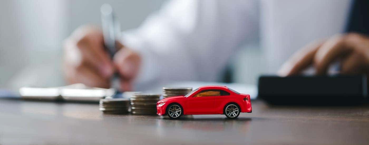 A graduating stack of coins is near a red toy car and person signing paperwork.