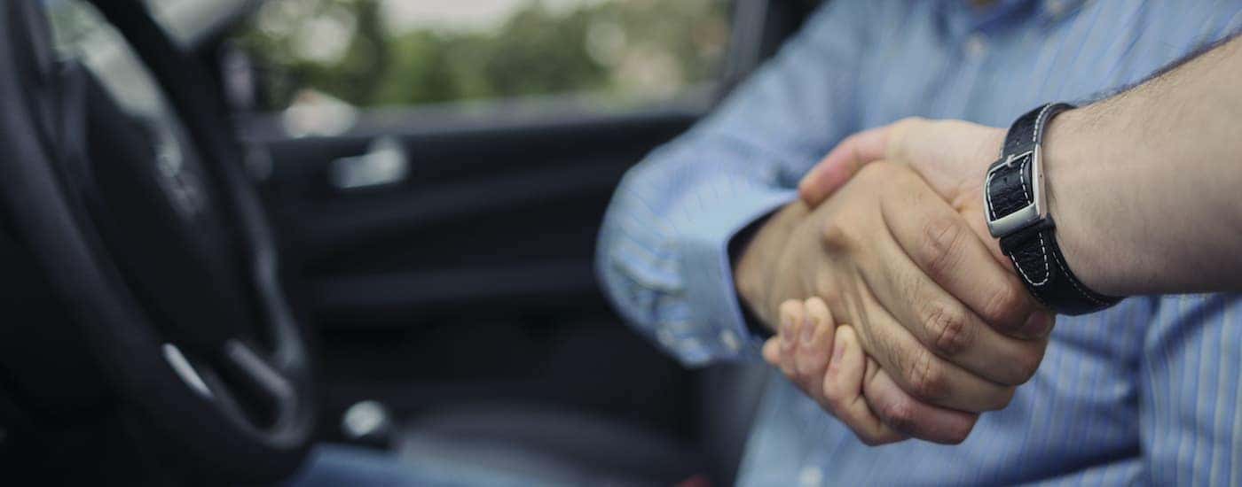 A person is shown sitting in the front seat of a car while shaking hands with a salesperson.
