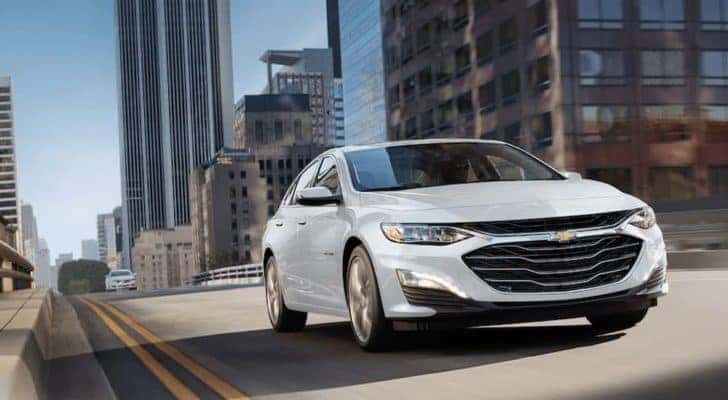 A white 2023 Chevy Malibu for sale is shown driving on a city street.