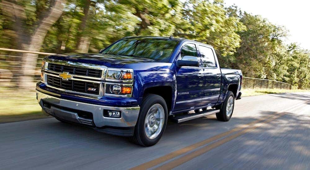 A blue 2014 Chevy Silverado 1500 LTZ Z71 is shown driving on a tree-lined road after a certified used truck dealer.