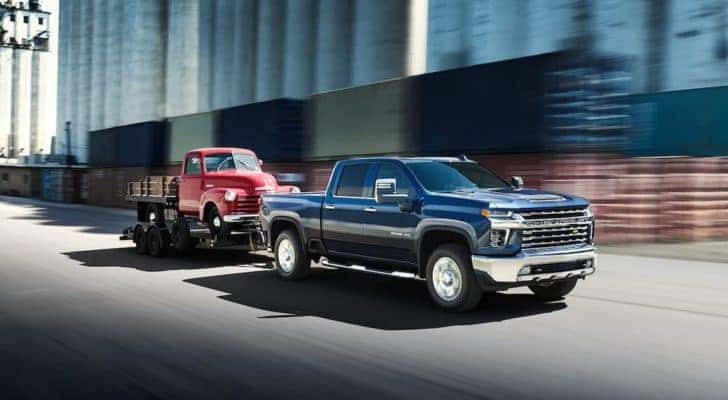 A blue 2022 Chevy Silverado 2500HD is shown towing a red truck on a city street.