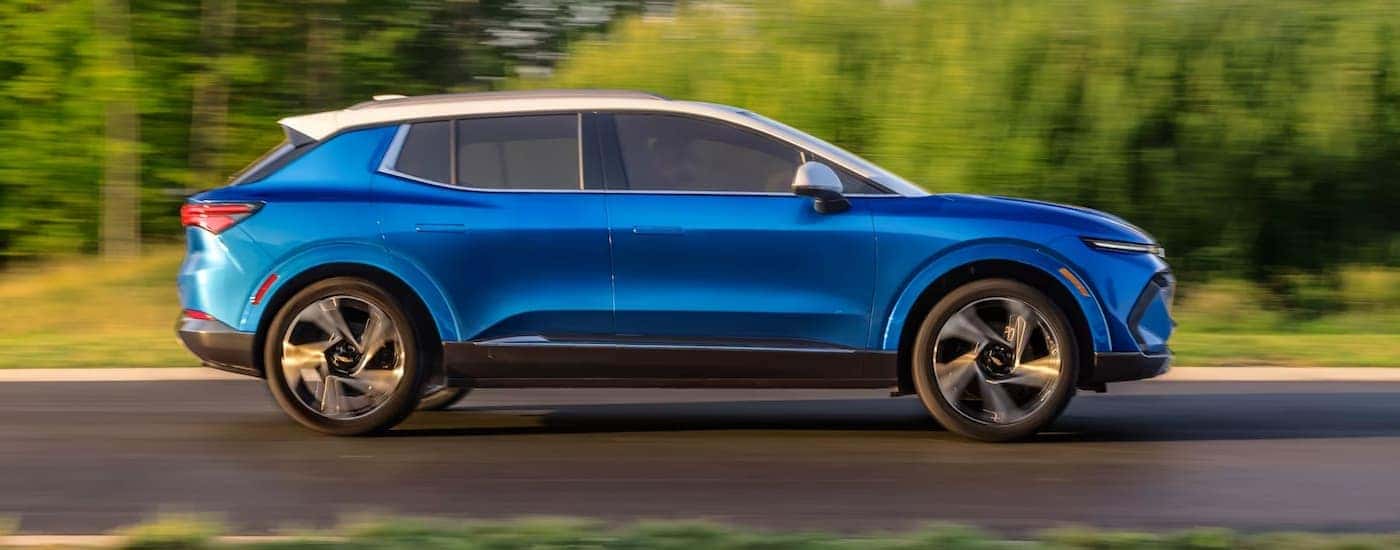 A blue 2024 Chevy Equinox EV is shown from the side driving on a tree-lined road.