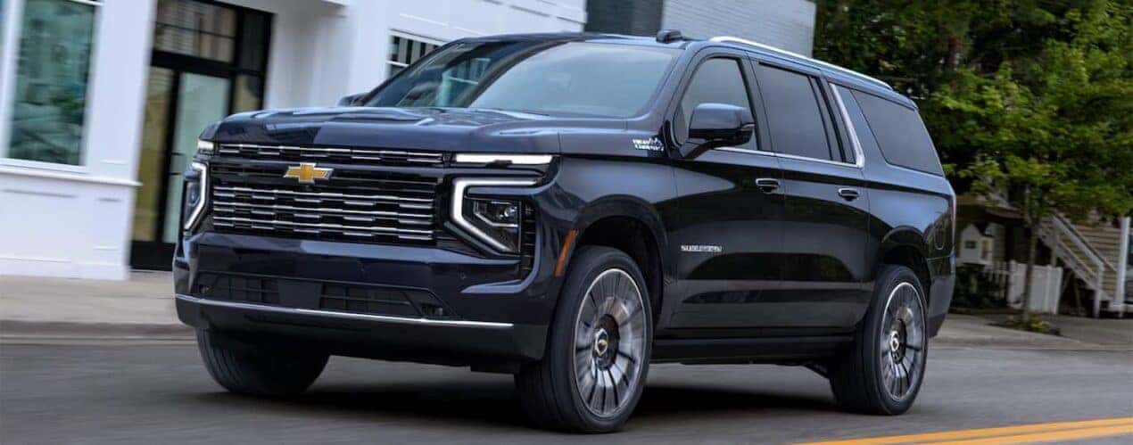 A dark blue 2025 Chevy Suburban is shown driving in a city.