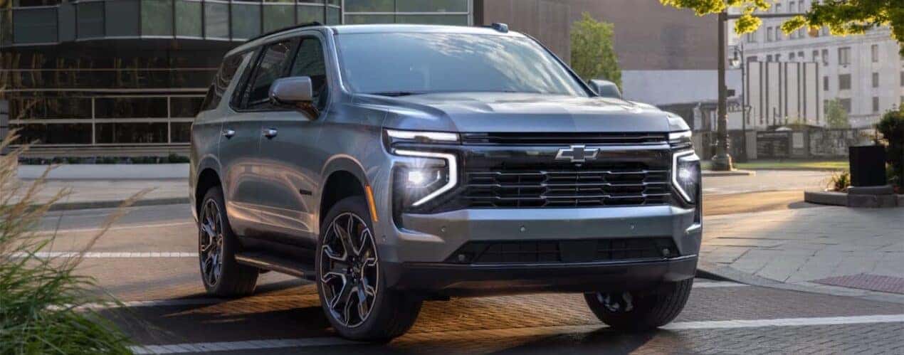 A grey 2025 Chevy Suburban is shown driving in a city.