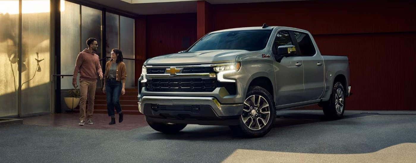 Two people are shown walking by a silver 2023 Chevy Silverado 1500 Z71 parked beside a building.