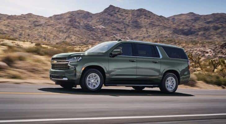 A sage green 2024 Chevy Suburban for sale driving on an open desert highway.