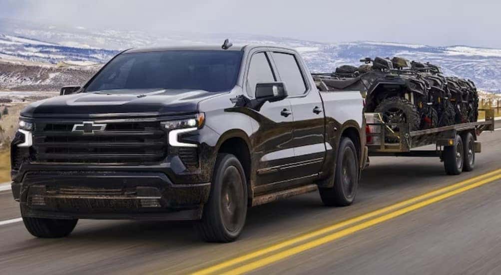 A black 2024 Chevy Silverado 1500 High Country is shown towing a trailer on a highway.