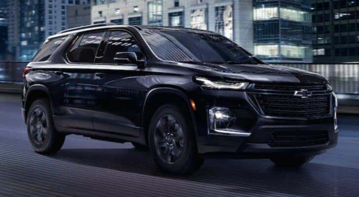 A popular used Chevy Traverse for sale, a black 2022 Chevy Traverse Midnight Edition, is shown driving on a city street.