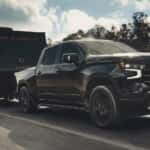 Black 2026 Chevy Silverado 1500 High Country towing a trailer down a road.