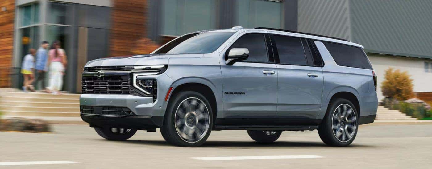 Silver 2026 Chevy Suburban driving past a modern apartment building.