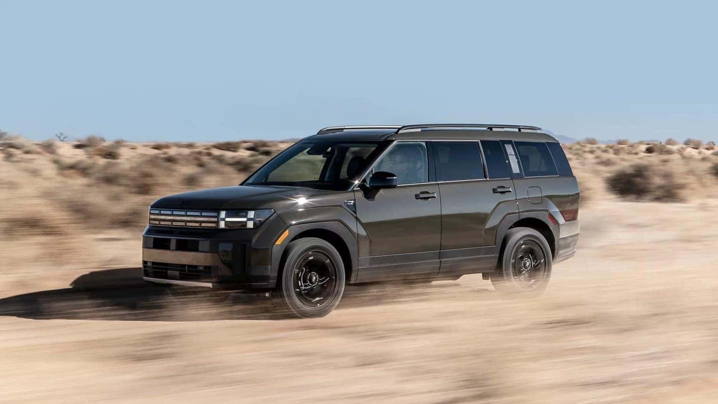 Dark gray Hyundai Santa Fe driving across a dusty desert road with low scrub and distant mountains.