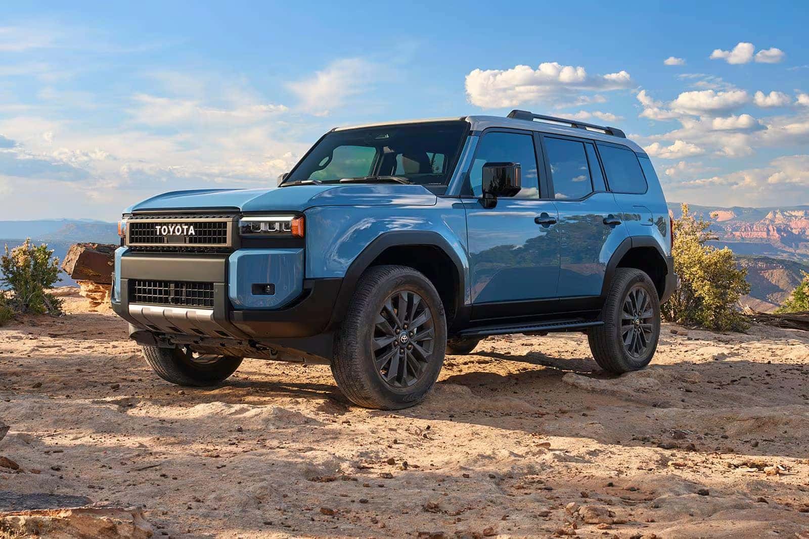 Blue Toyota Land Cruiser parked on a rocky overlook with canyon scenery and a bright blue sky.