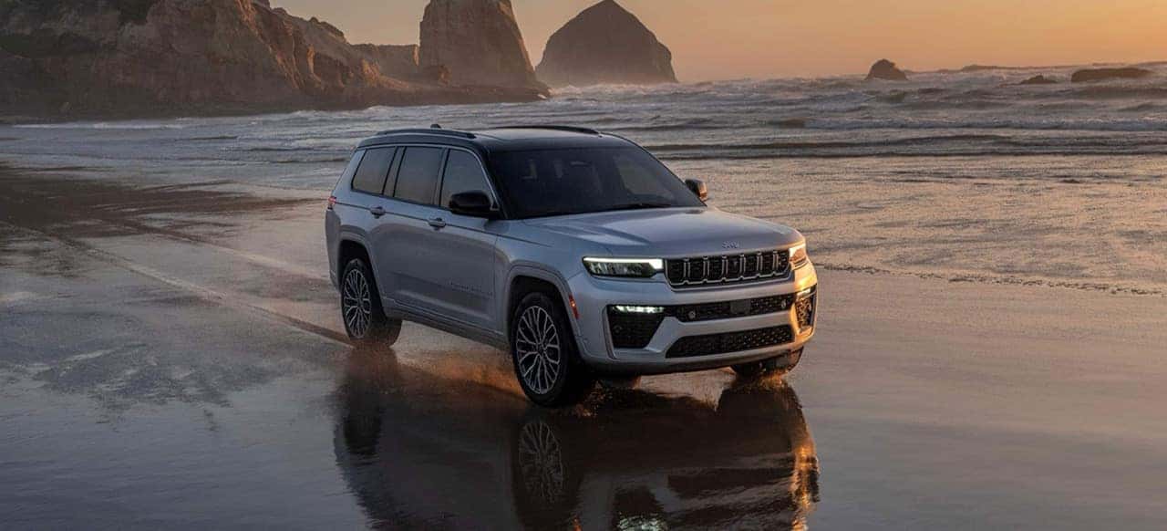 Silver Jeep Grand Cherokee driving across wet sand on a beach at sunset with ocean waves and sea stacks behind it.