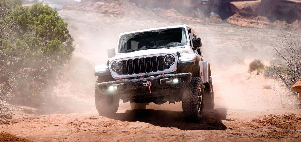 White Jeep Wrangler driving over a rocky desert trail with dust rising around the vehicle.