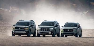 three dark colored TrailSport SUVs drive down a dirt road, the dust they kick up is obscuring the mountains behind them