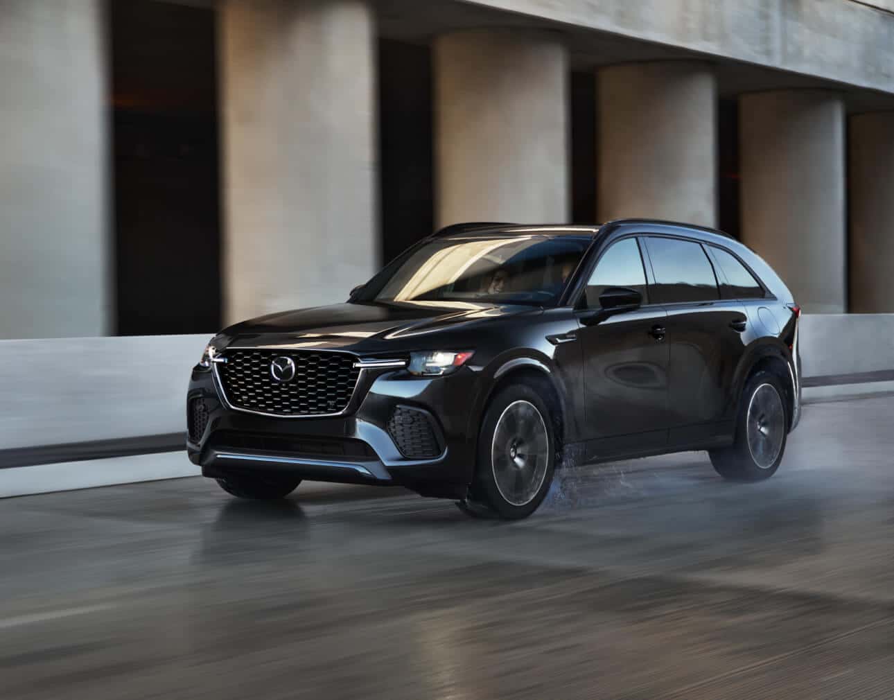 Front three-quarter motion view of a black 2026 Mazda CX-70 driving past concrete pillars.