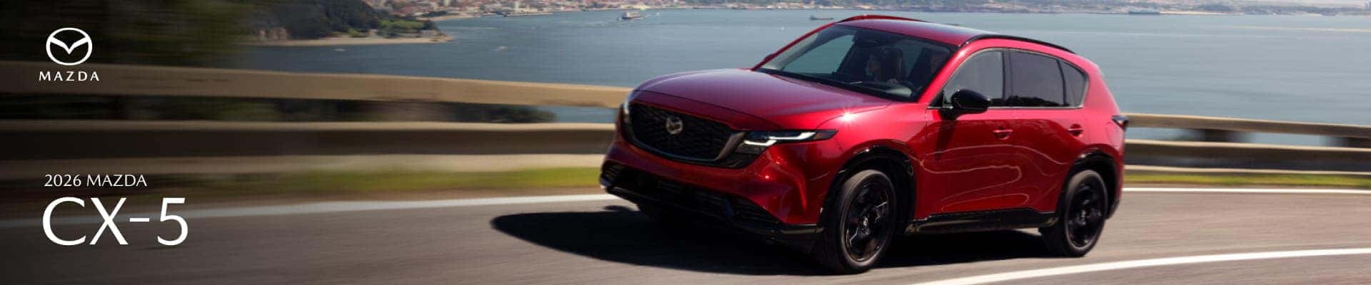 Red 2026 Mazda CX-5 driving along a coastal road, shown from the front three-quarter view.