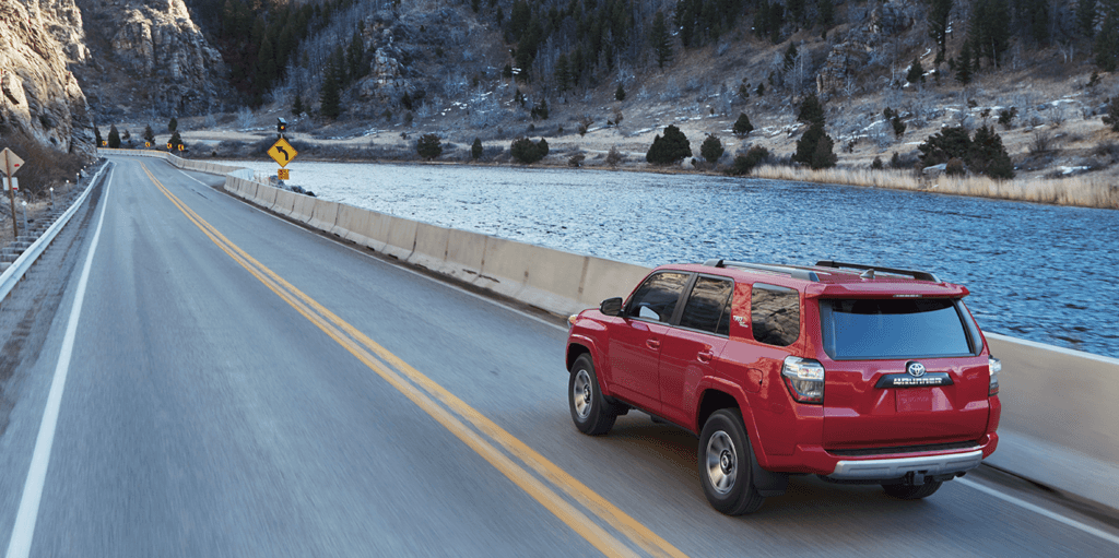 What is Tire and Rim Protection? | Lancaster Toyota | Rear view of a Red Toyota 4Runner driving down road with lake on the right side 