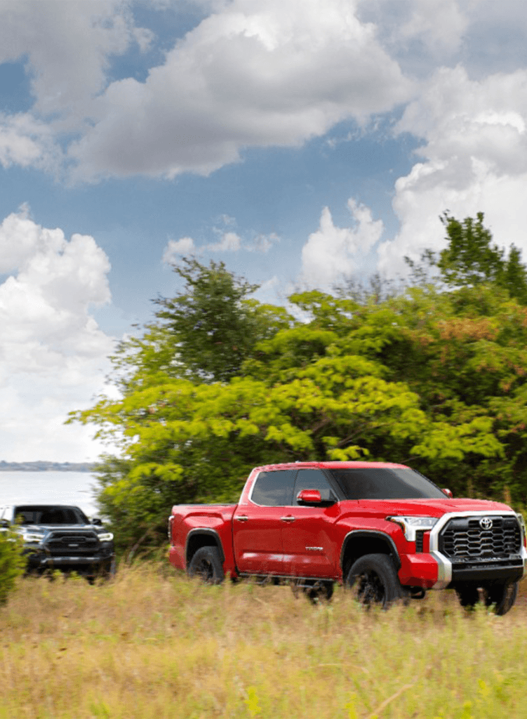 2023 Tundra vs 2023 Tacoma at Lancaster Toyota | Red Toyota Tundra driving through some tall grass while a dark color Toyota Tacoma is right behind it