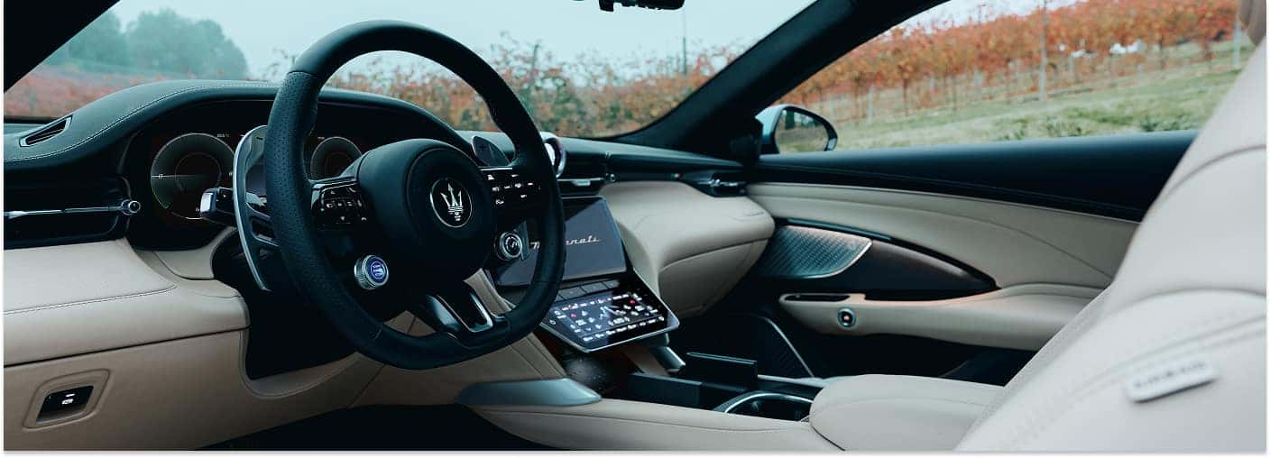The luxurious interior of a 2026 Maserati GranTurismo features a black leather-wrapped steering wheel with the prominent trident emblem, large silver paddle shifters, dual touchscreen center displays, and premium cream-colored leather upholstery.