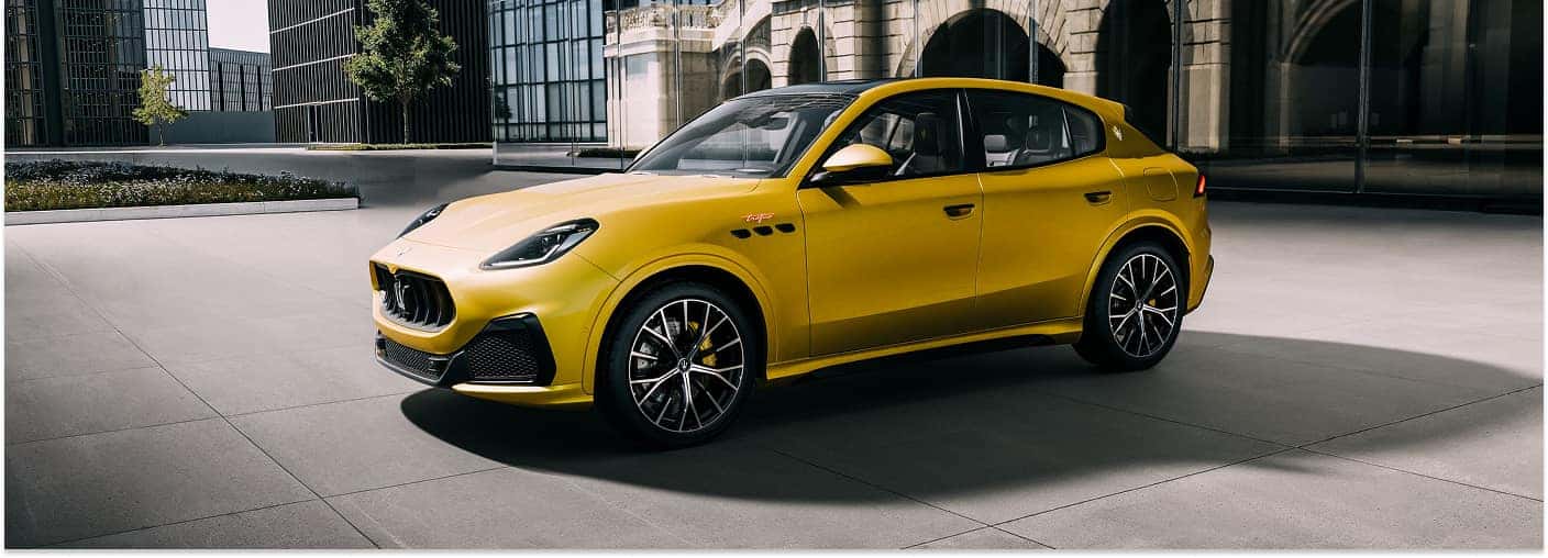 A front three-quarter view of a bright yellow 2026 Maserati Grecale Trofeo parked on a paved urban plaza with modern and arched buildings in the background.